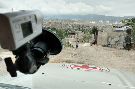 View out the windshield of a car on a dirty road overlooking a sprawling beige city. Visible is a Red Cross symbol on the car hood and a dash camera mounted to the windshield.